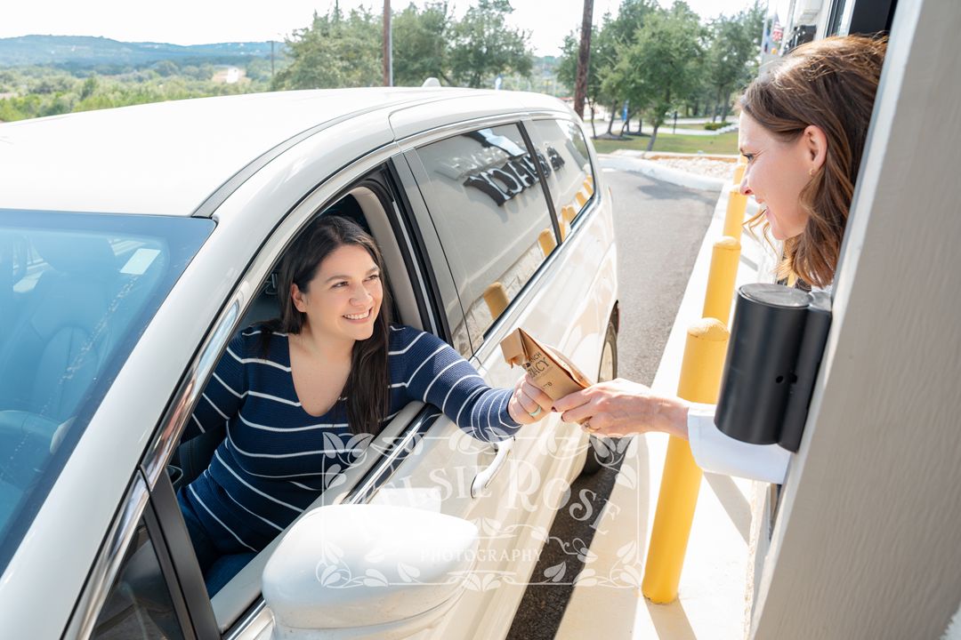 Woman in car receiving prescription bag from pharmacy drive-through window attendant on a sunny day.