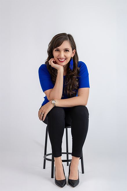 Woman in blue top and black pants sitting on stool smiling confidently in professional studio portrait.