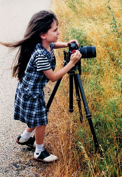 Young girl with long brunette hair wearing plaid using DSLR camera on tripod outdoors in field.