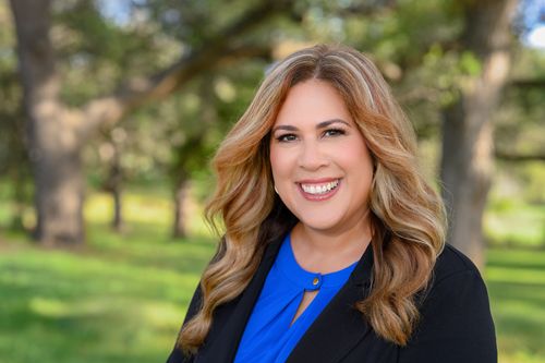 Professional headshot of businesswoman with brunette hair smiling outdoors, wearing a blue top and black blazer, with trees in background.