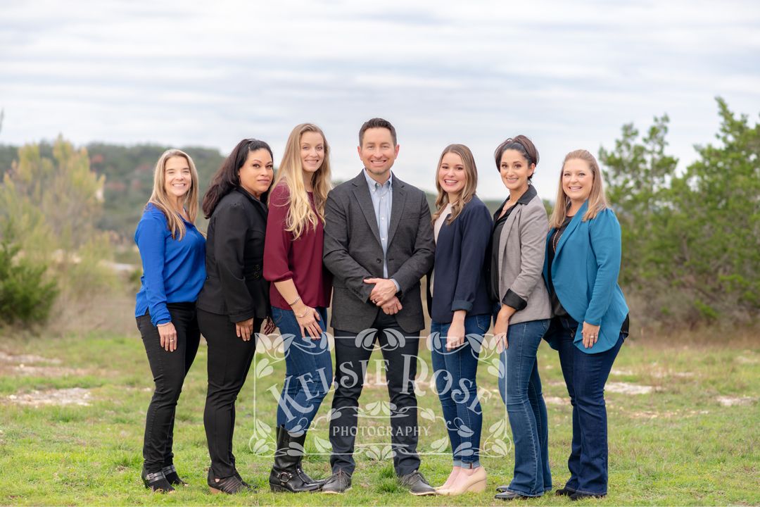 A dental team of seven professionals poses outdoors in casual attire on a cloudy day.