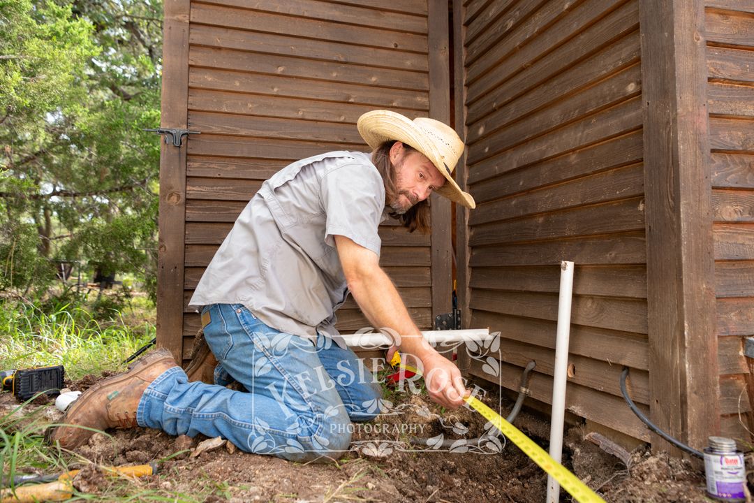 Worker in cowboy hat kneeling to measure ditch along the base of a wooden well building.