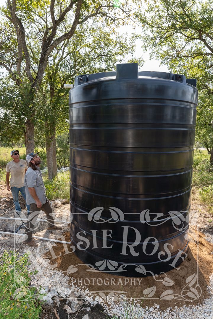 Two male workers looking up at a large black water storage tank outdoors among trees on a sunny day.