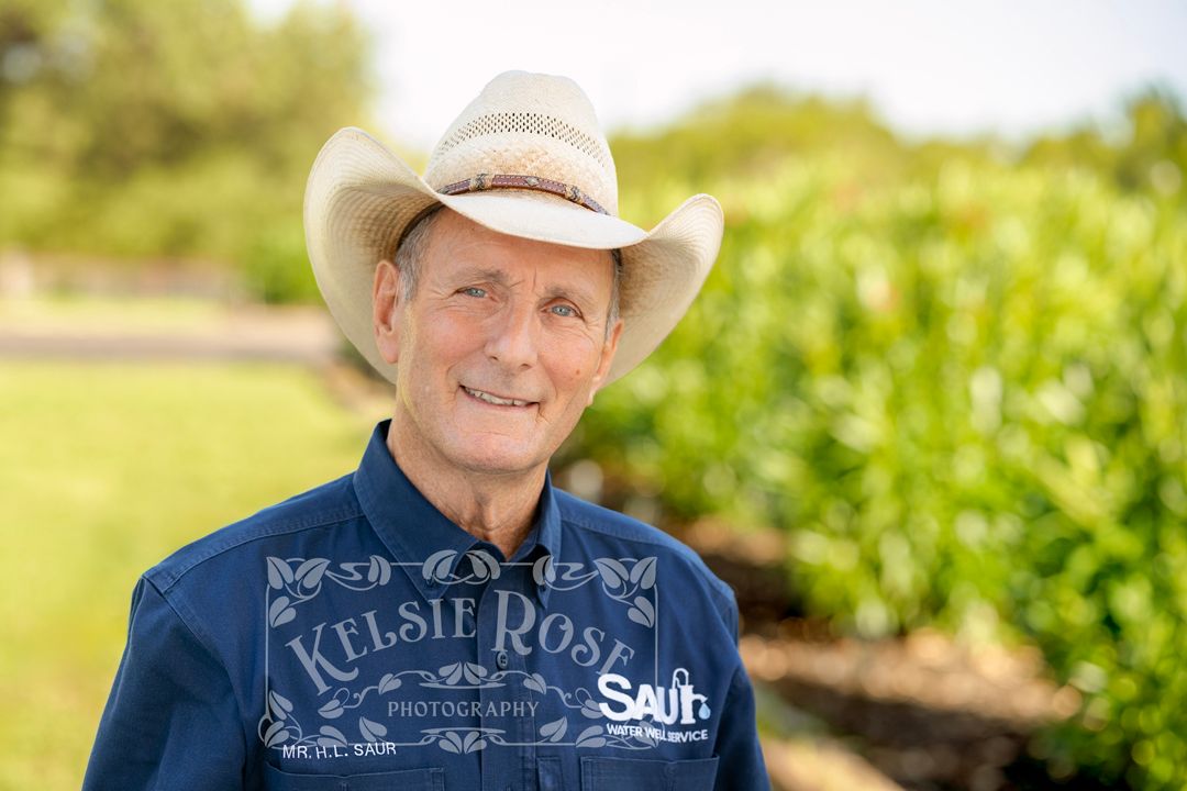 Smiling older man in white cowboy hat and blue denim shirt outdoors with oleander bushes in background in a rural Texas setting.