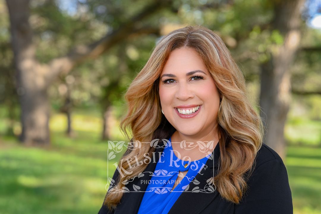 Professional headshot of a smiling woman with brunette hair wearing a blue top and black blazer outdoors.
