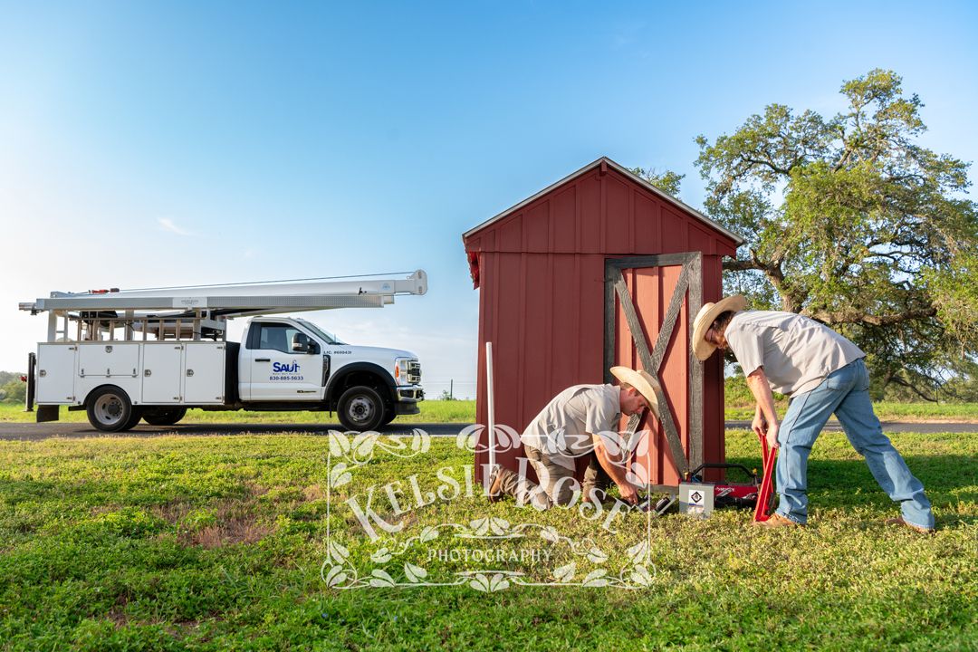 Two technicians working at base of red well building with well service truck in background.