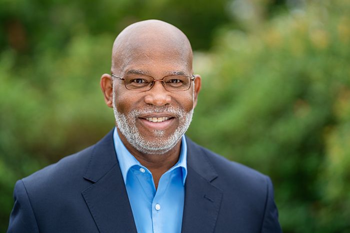 Professional headshot of a smiling middle-aged man in a blue shirt and navy blazer outdoors.