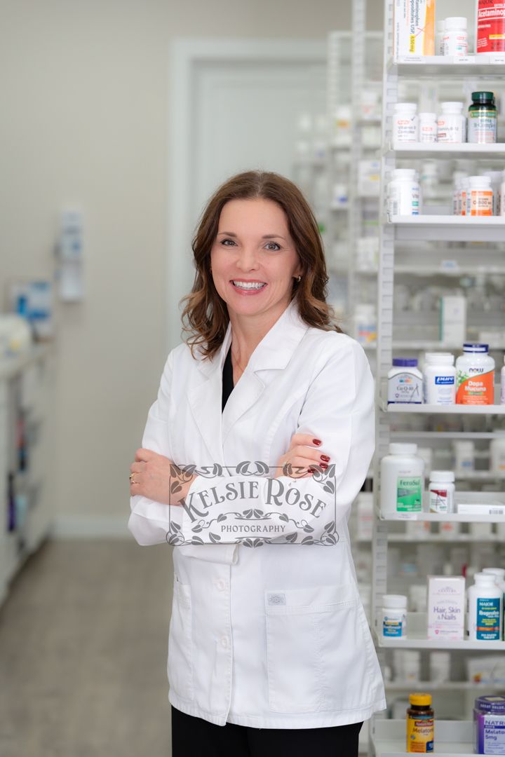 Female pharmacist in white lab coat smiling with arms crossed beside shelves of health products.