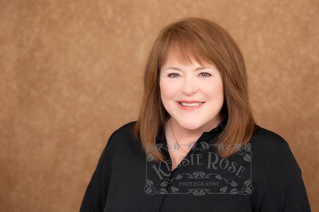 Professional headshot of smiling woman with auburn hair wearing black top against brown background.