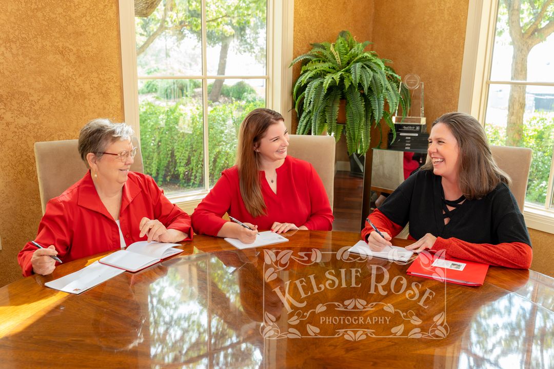 Three women in red tops smile and review documents at a polished wooden table in an elegant room.