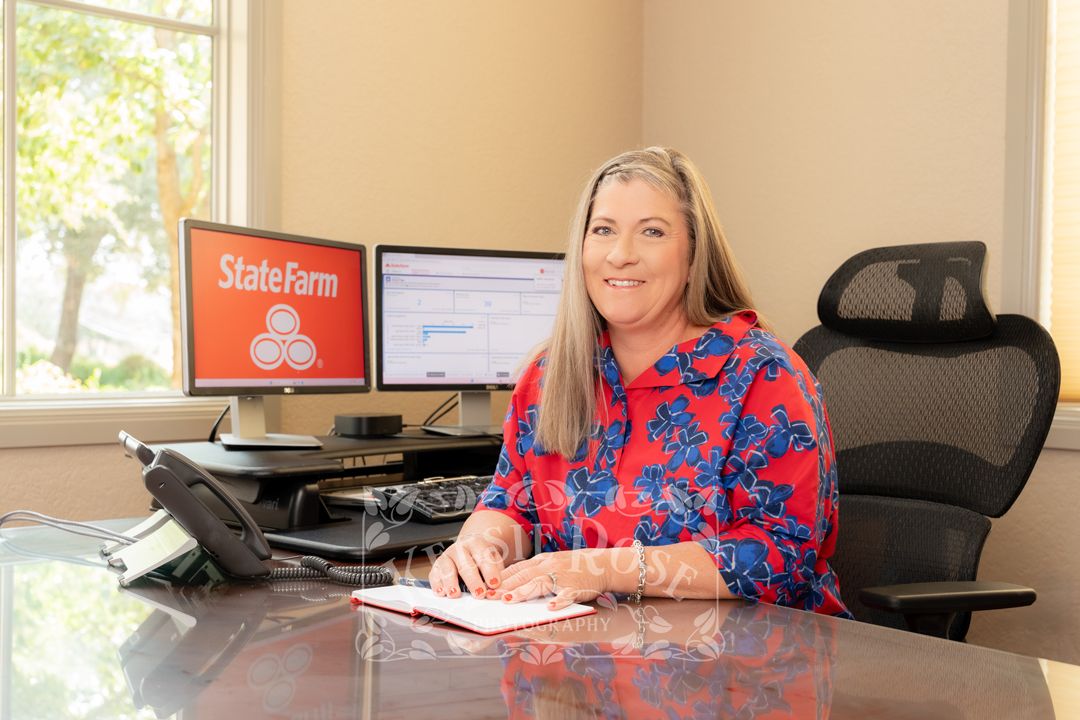 State Farm agent smiling at desk with dual monitors displaying State Farm logo in a professional office setting.