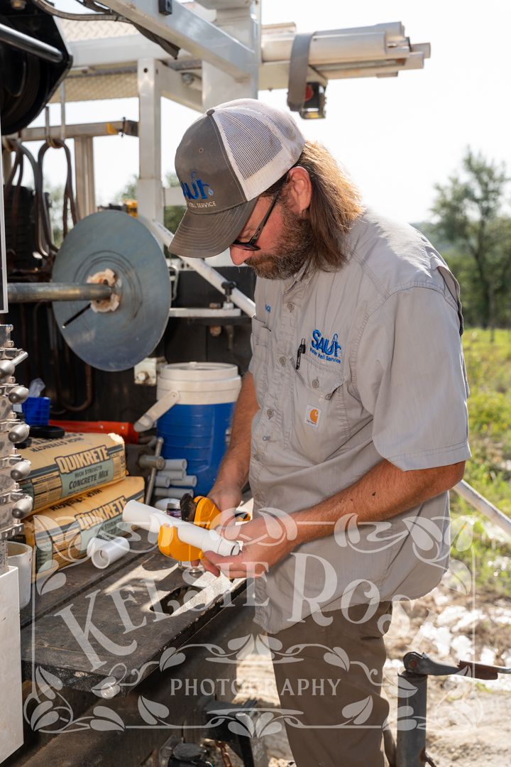 Bearded man in cap and work shirt applies caulk to PVC pipe outdoors near industrial equipment on a sunny day.