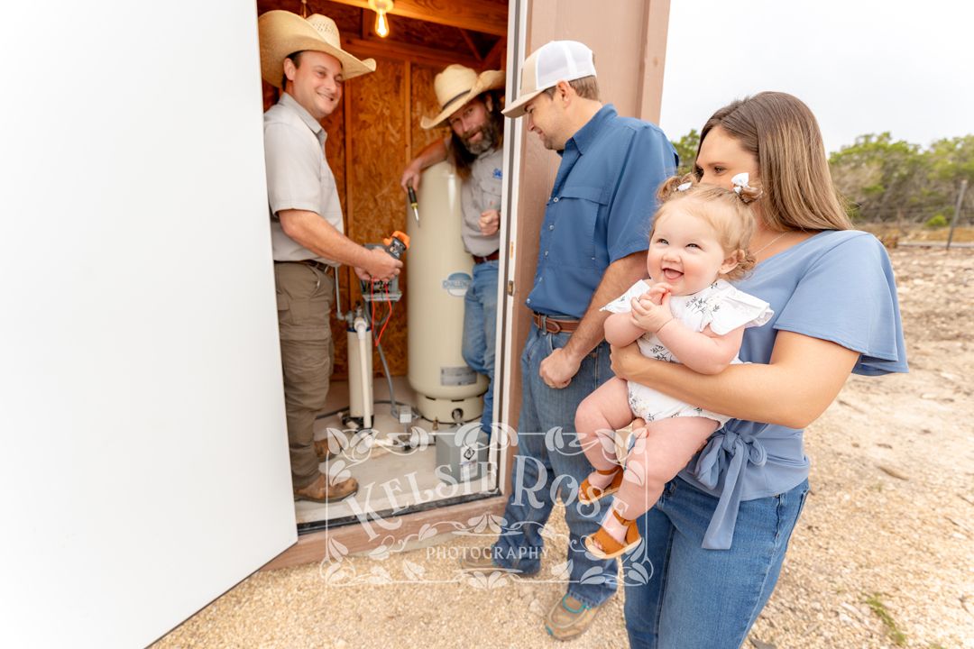 Water well service technicians installing equipment while a family with a baby watches outside a well building in Texas.