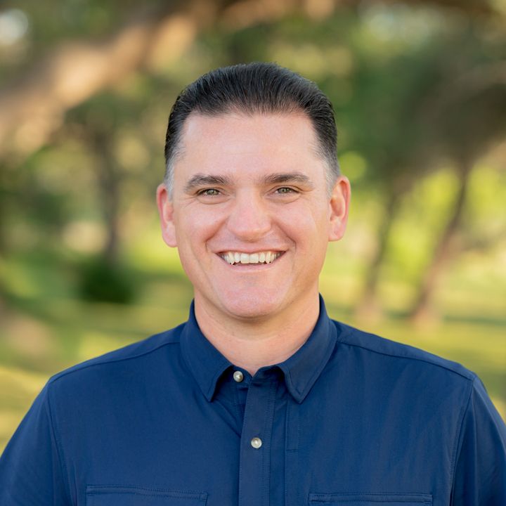 Cheerful man wearing navy shirt smiles warmly in outdoor professional portrait with green trees behind him.