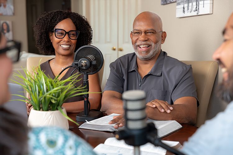 A smiling couple records a podcast together at a table with microphones and an open Bible, interviewing a second couple.