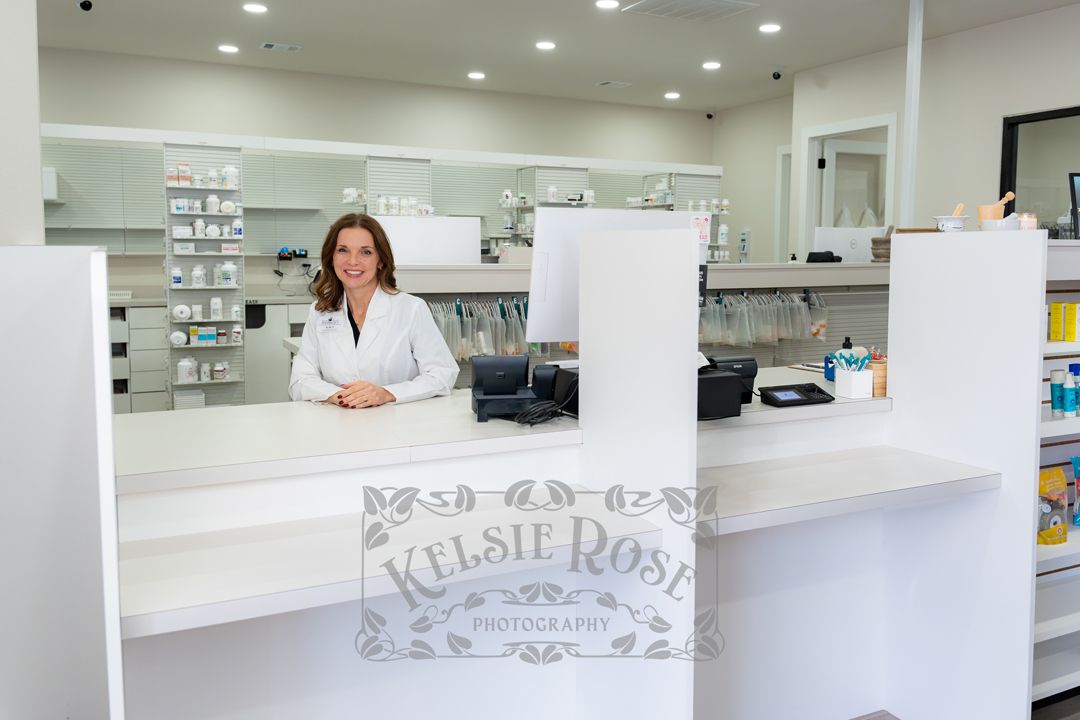 Female pharmacist in white lab coat standing behind a bright white modern pharmacy dispensing counter.