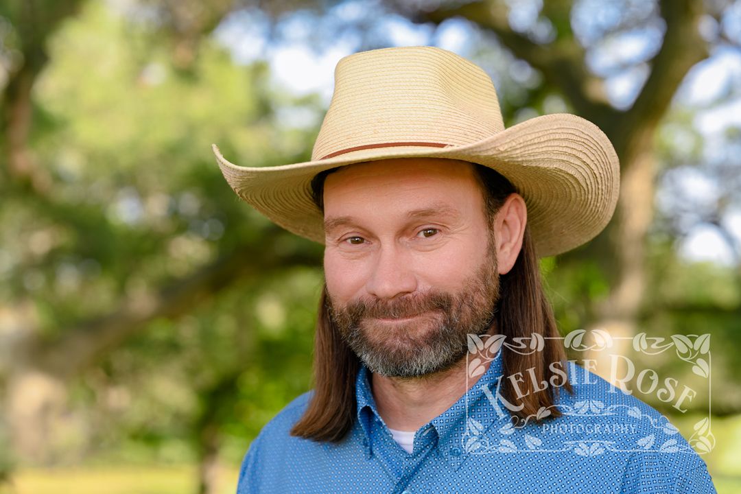 Headshot of man smiling in straw cowboy hat with long hair and beard outdoors.