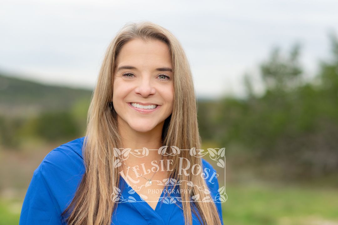 Headshot of smiling woman with long brown hair in blue shirt outdoors with Texas Hill Country in background.