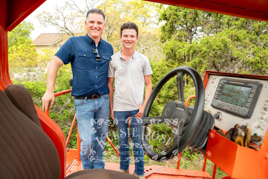 Father and son smiling together next to the cab of an orange road paving machine.