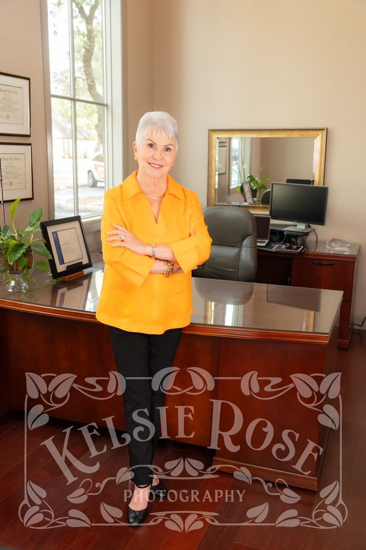 Professional woman in orange blouse stands confidently with arms crossed in front of an executive desk.