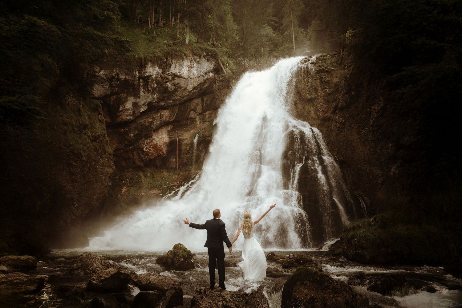 Svadobná fotka nevesty a ženícha, ktorí stoja pred majestnátnym vodopádom Gollingerwasserfall v moody atmosfére.
