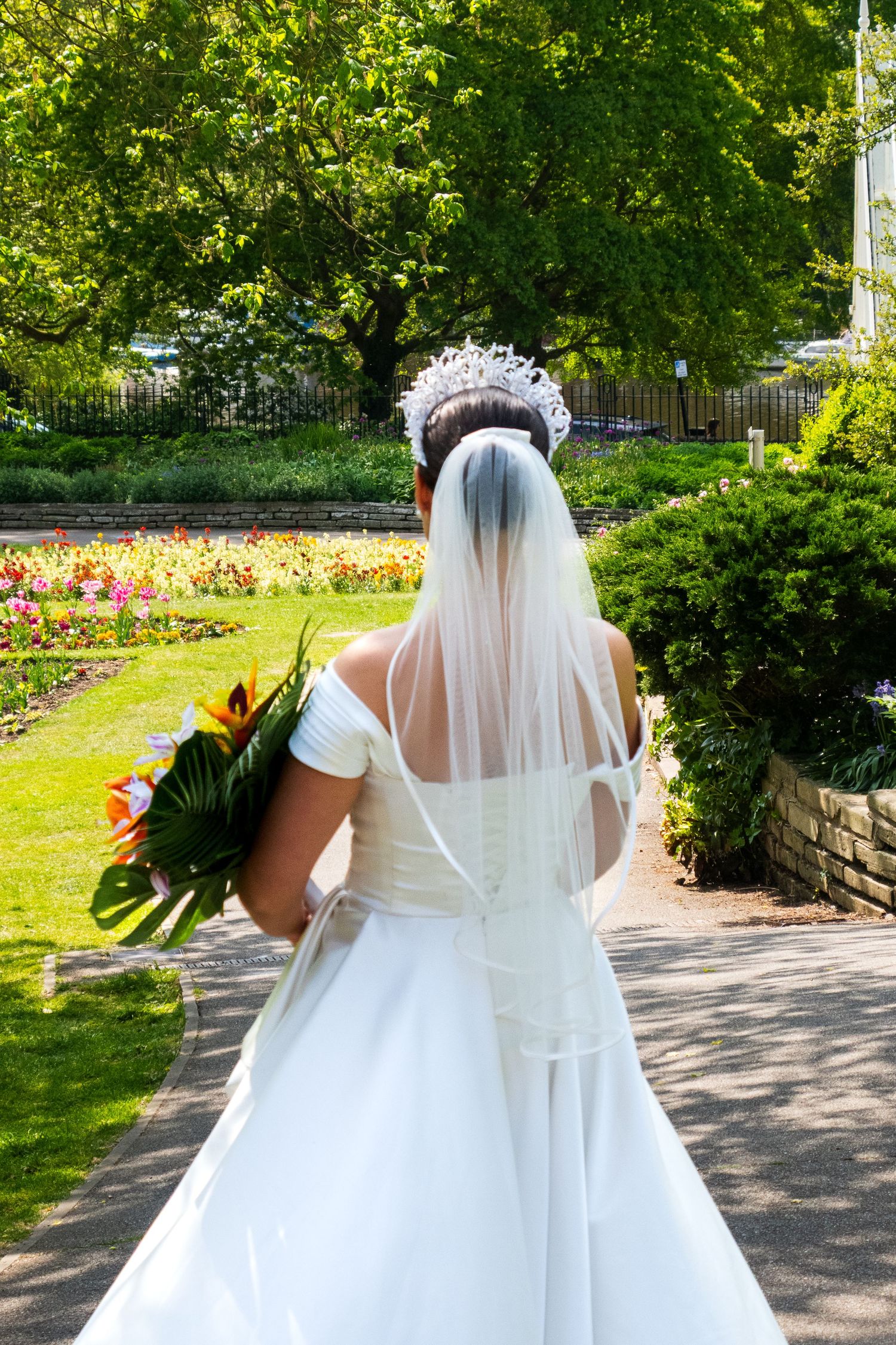 Back view of a bride in a white gown and crystal crown walking through a garden path.