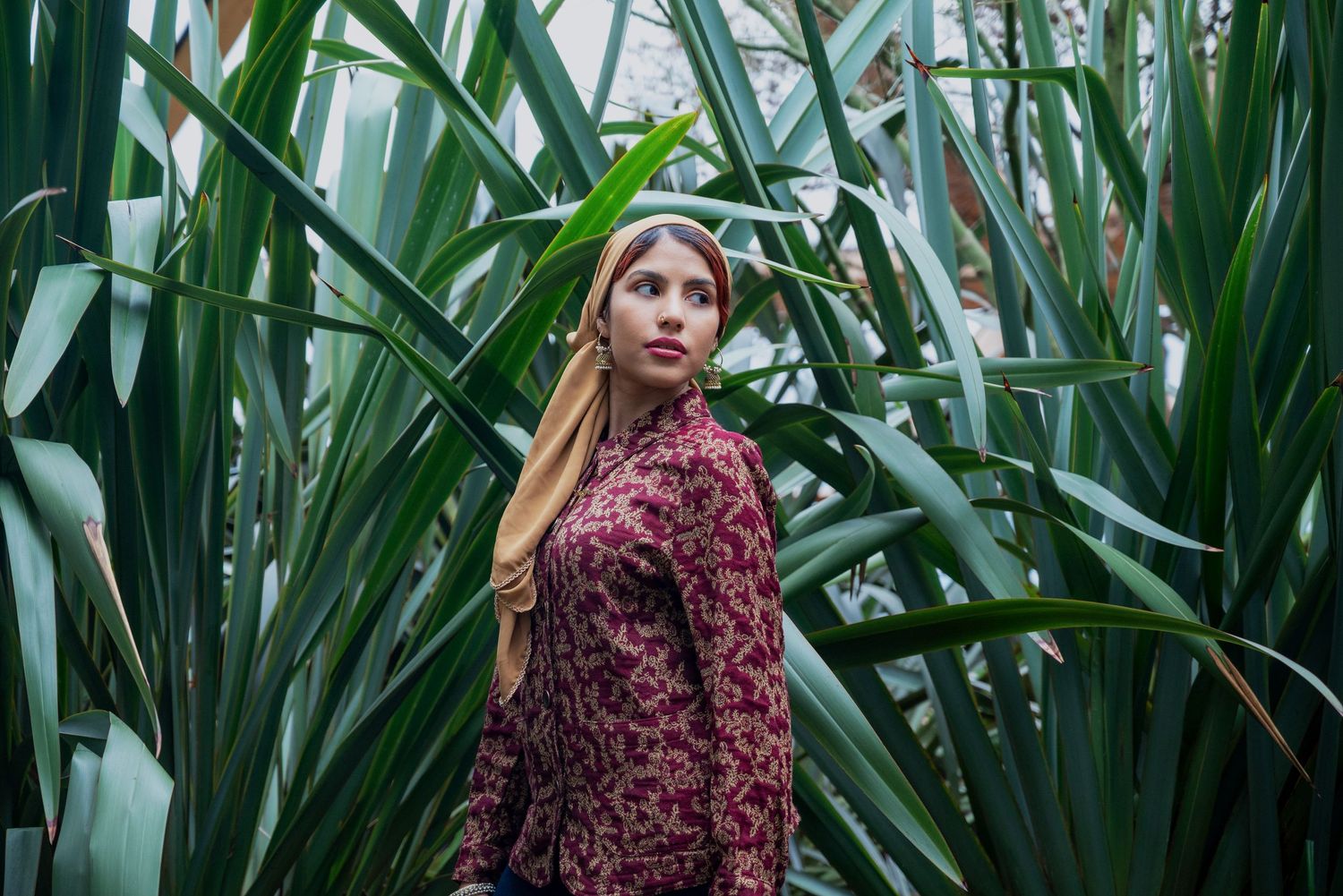 Young woman in floral dress standing among tall green tropical plants, looking over her shoulder.