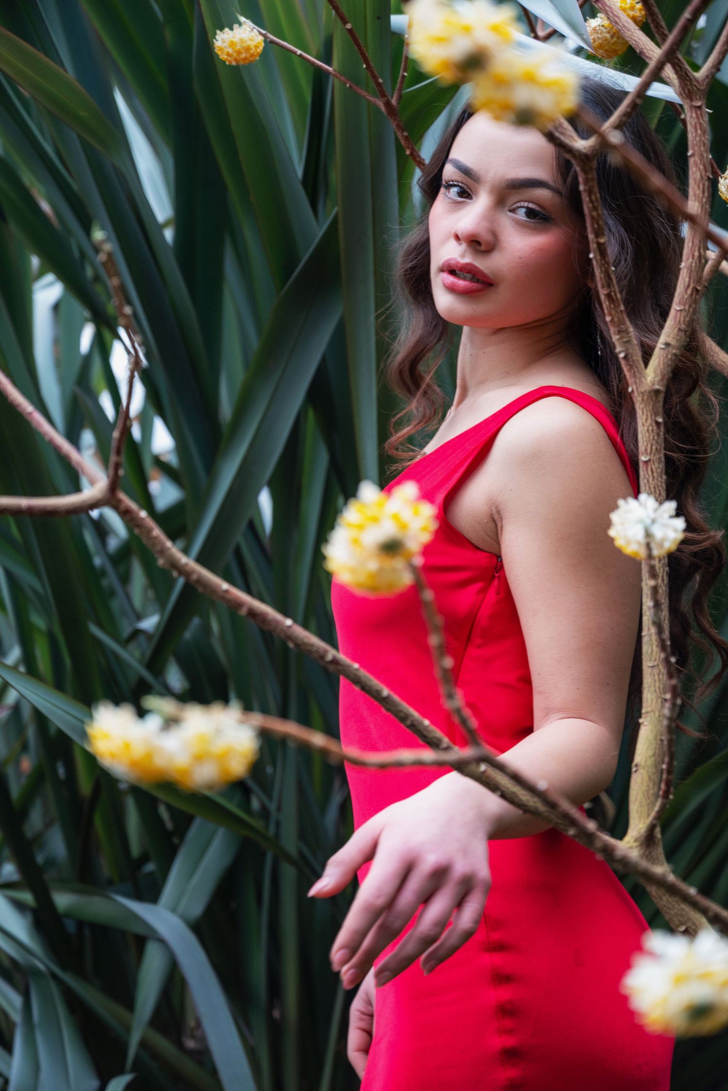 Young woman in red dress posing among tropical plants with yellow flowers in a lush green garden.