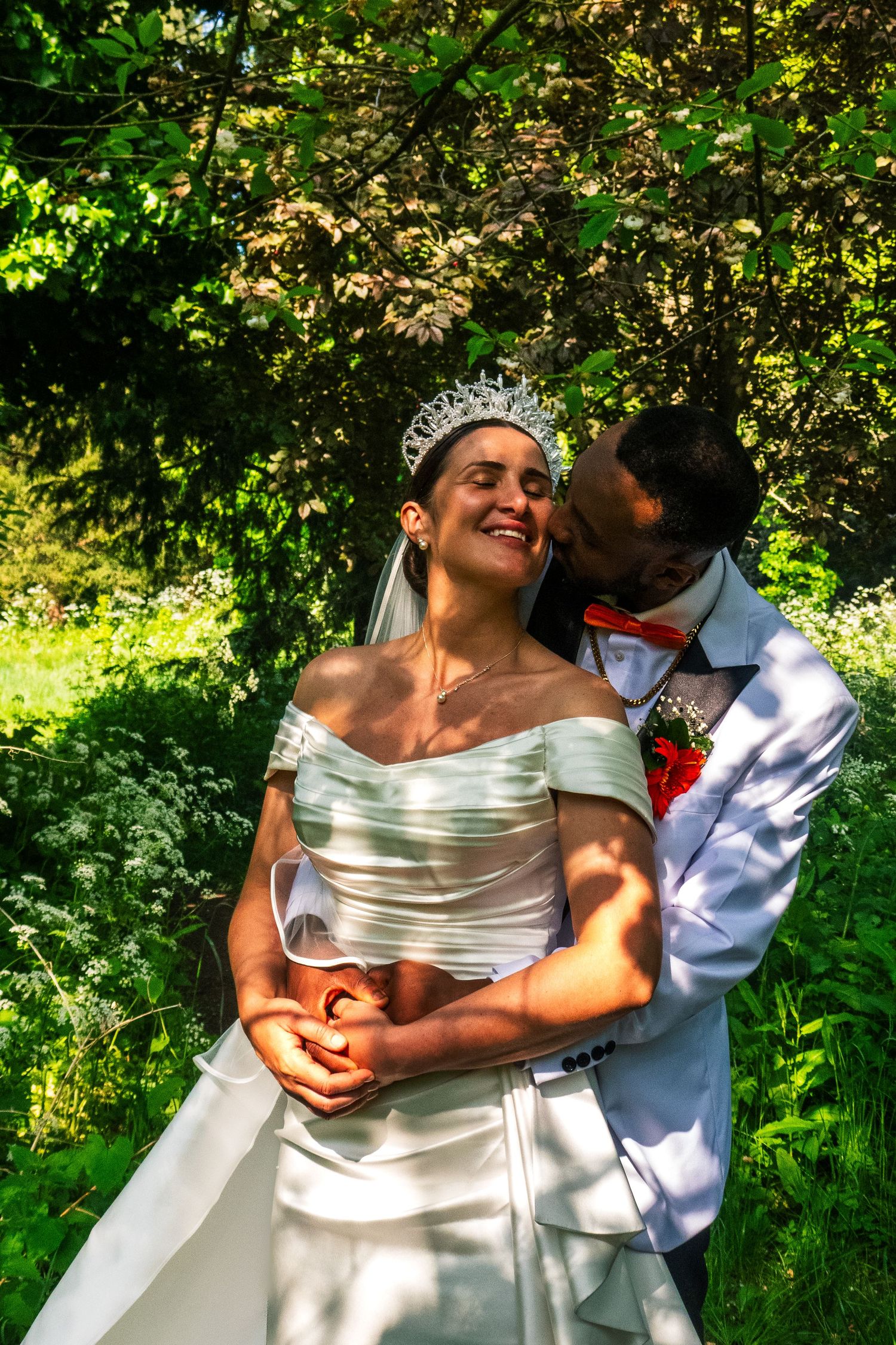 A candid moment between newlyweds sharing laughter in a sun-dappled garden setting.