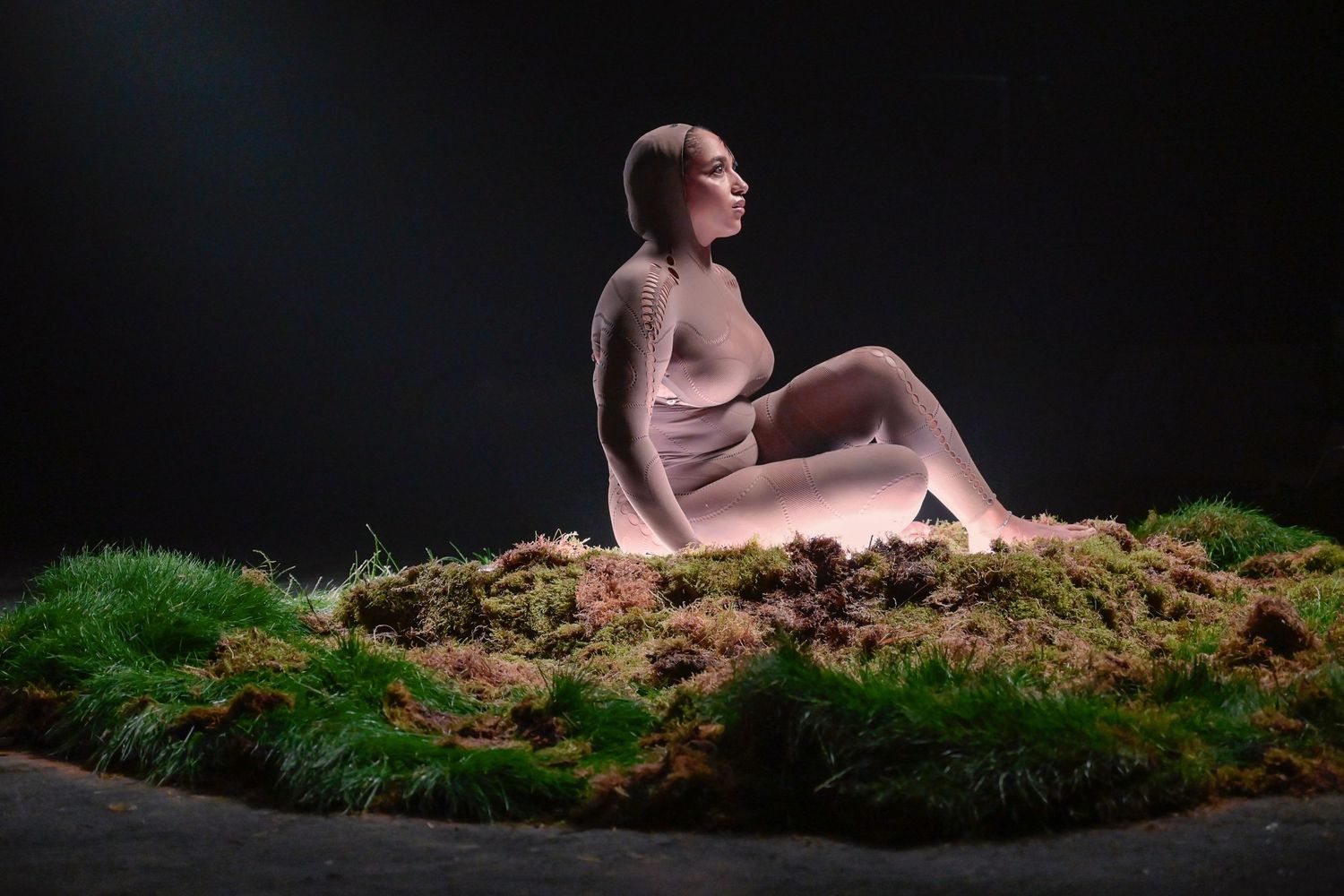 Artistic sculpture figure seated on mossy surface against dark background.