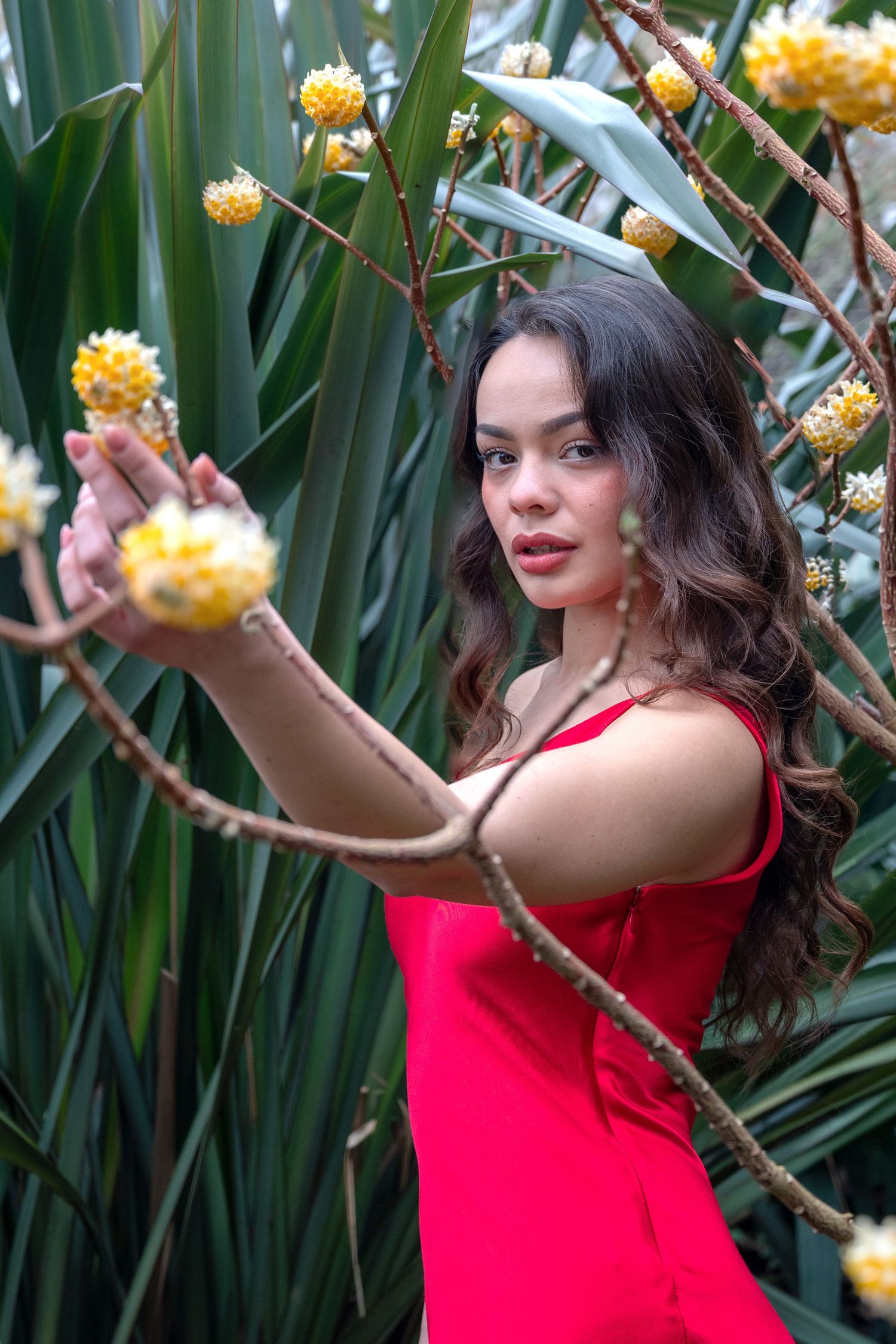 Young woman in red dress touching yellow flowers among tropical green plants.