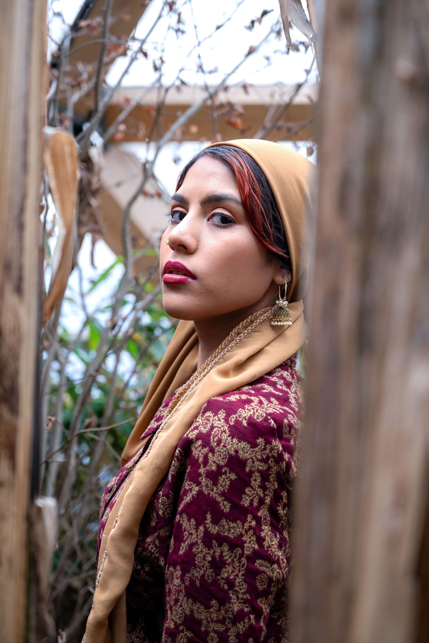 A young woman in a gold hijab and burgundy floral jacket poses outdoors near wooden beams and bare branches.