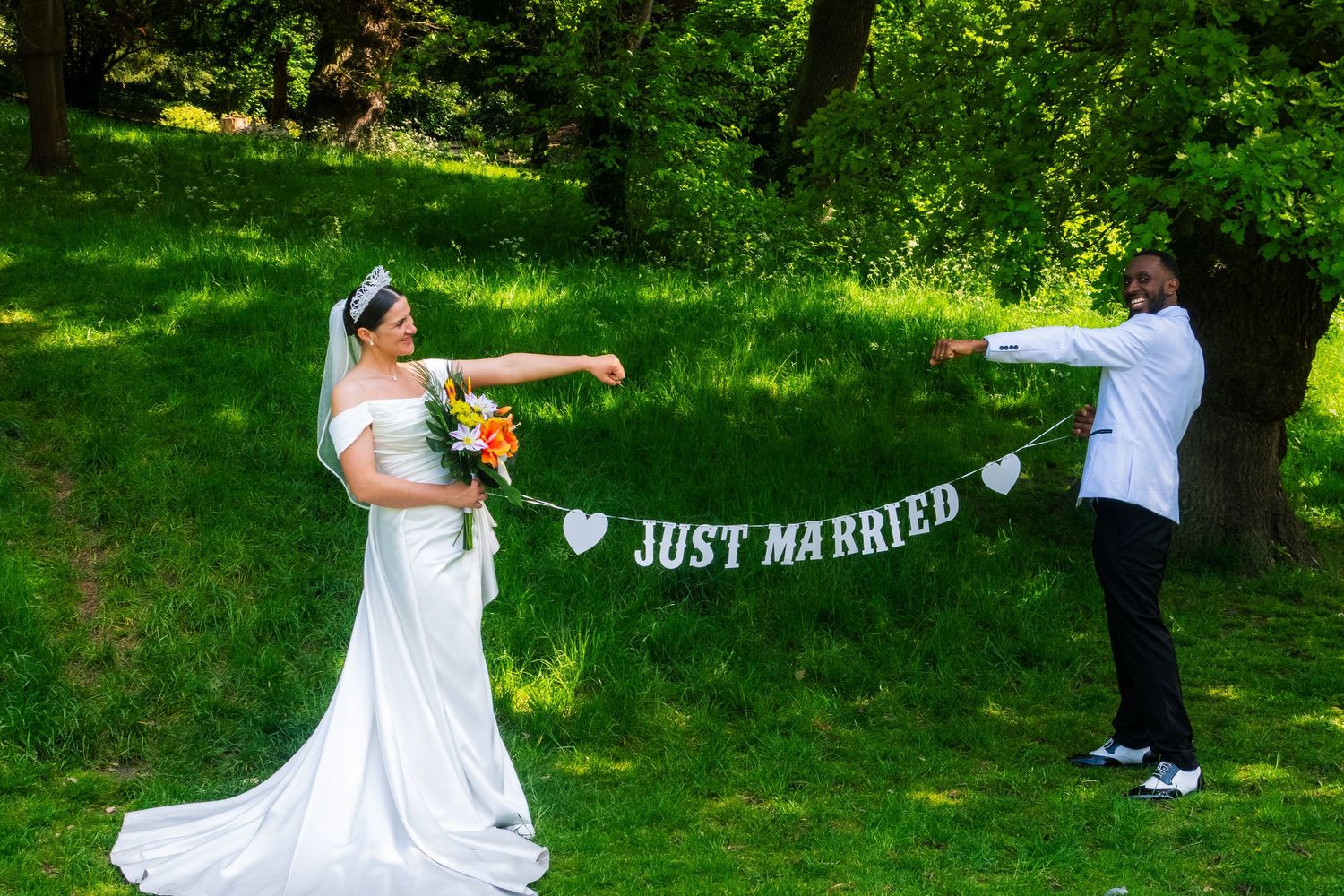 A couple poses playfully with a 'Just Married' banner in a sunny garden setting.