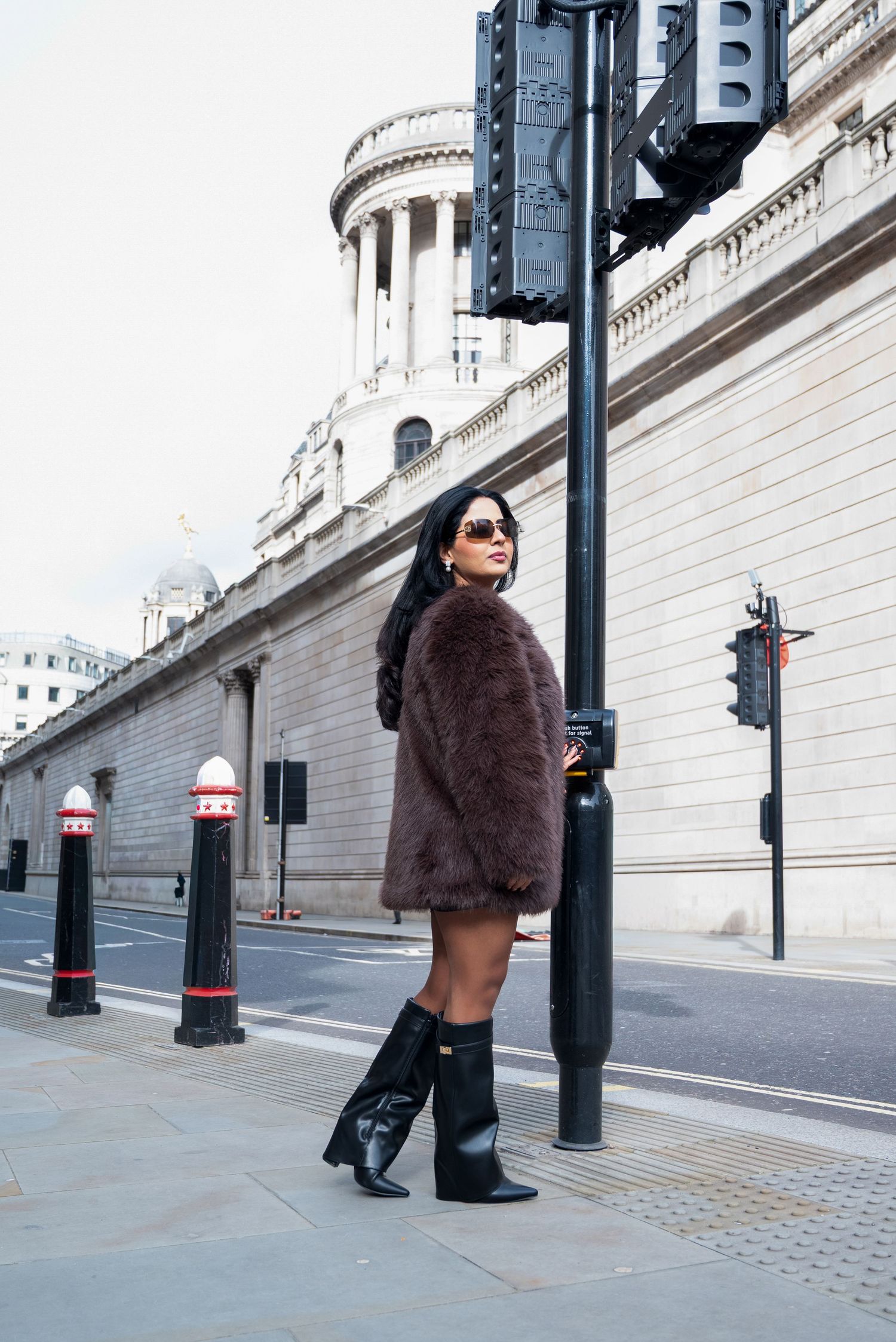 Person in brown fur coat and sunglasses walking past ornate stone building with decorative columns and lamp posts.
