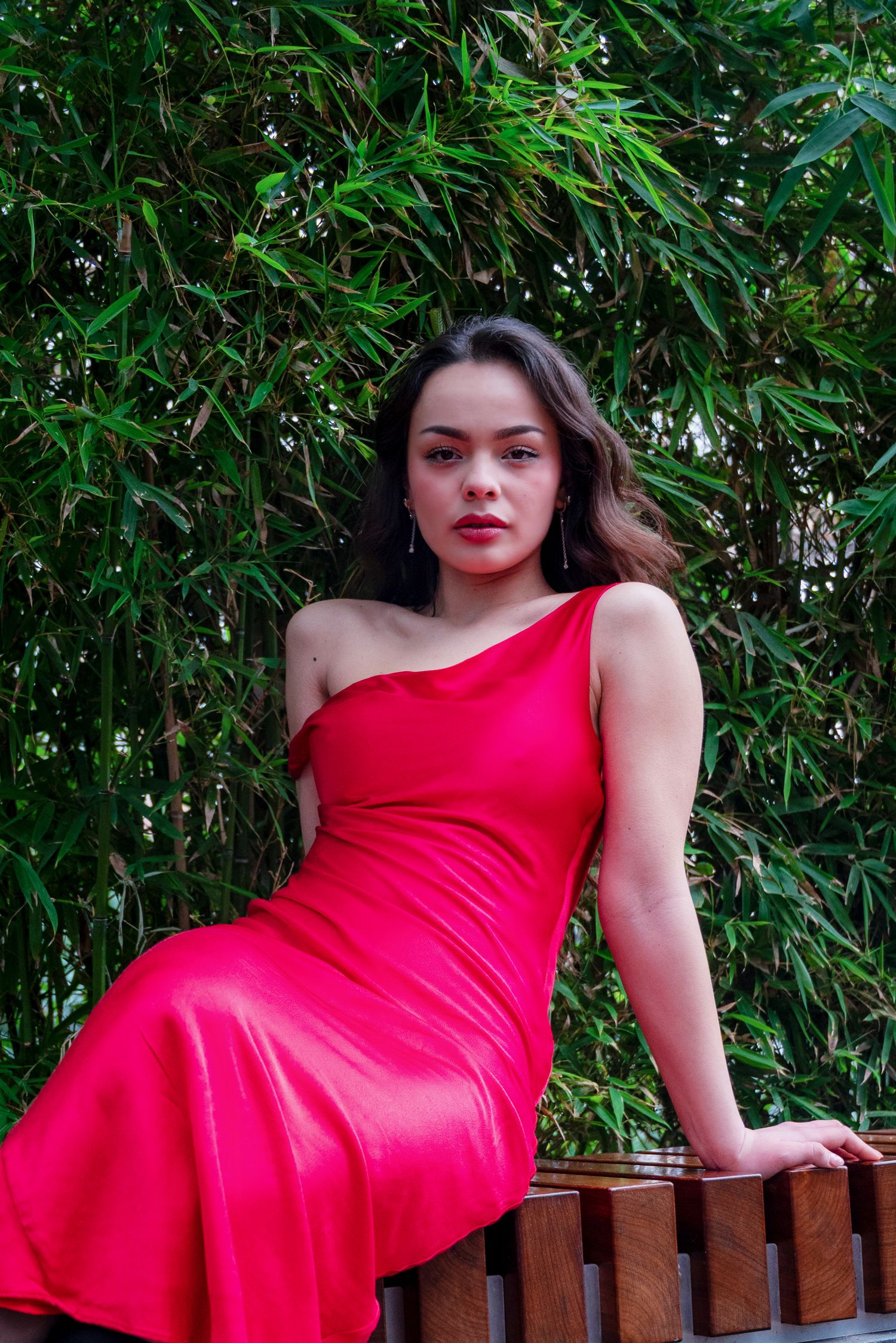 Young woman in elegant red one-shoulder satin dress seated on wooden bench against lush green bamboo backdrop.