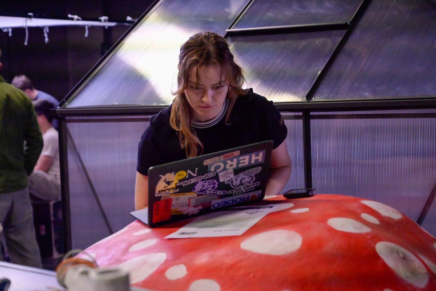 Person works on laptop while sitting next to large red and white spotted mushroom prop.