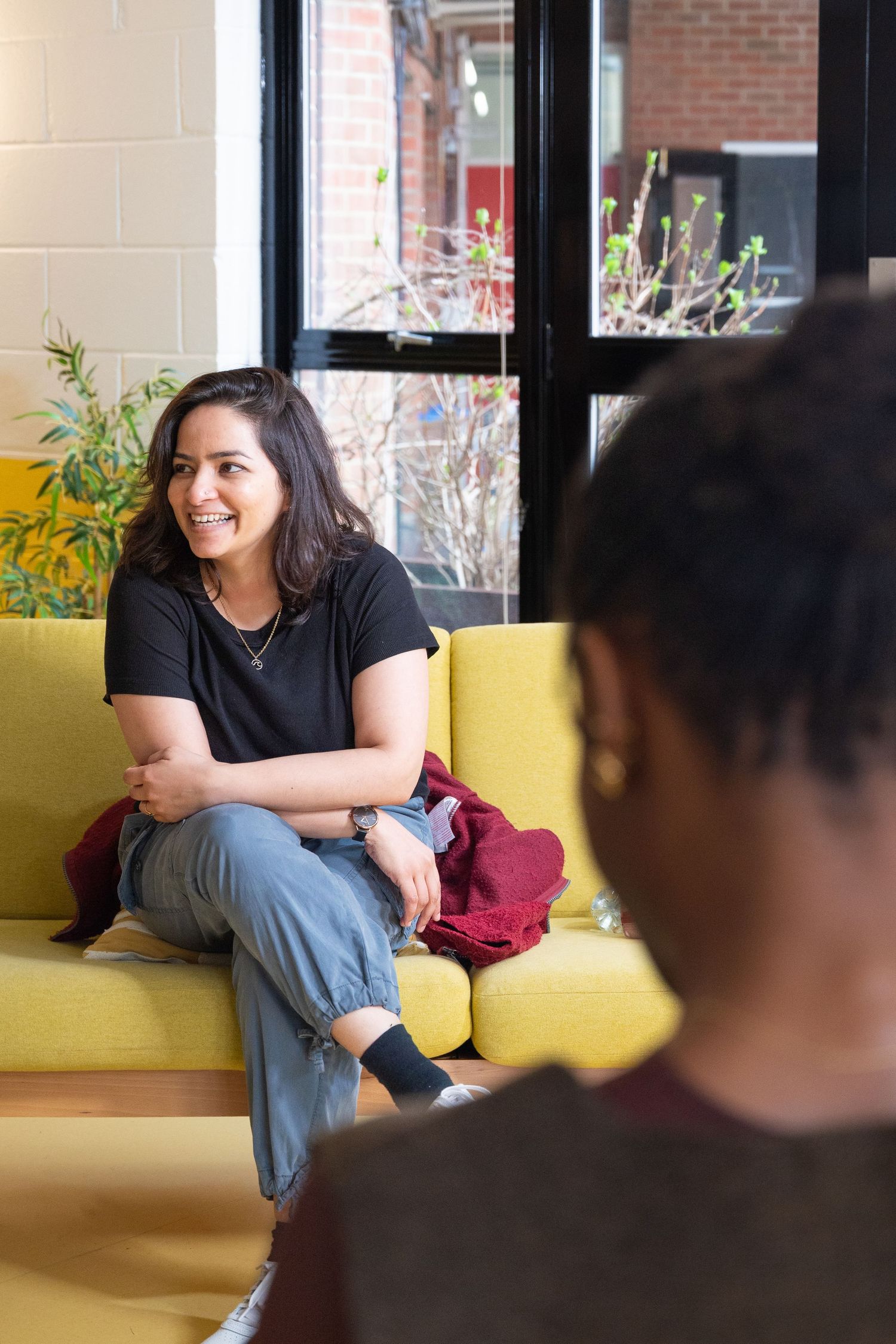 Person wearing black shirt and jeans sits casually on yellow couch in sunlit room with brick wall.