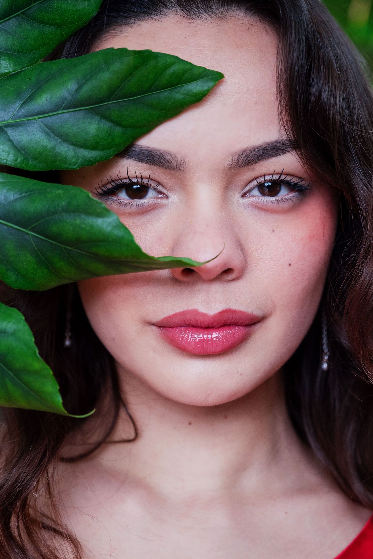 Young woman with brown eyes and rosy lips poses behind large green tropical leaves in a natural beauty portrait.