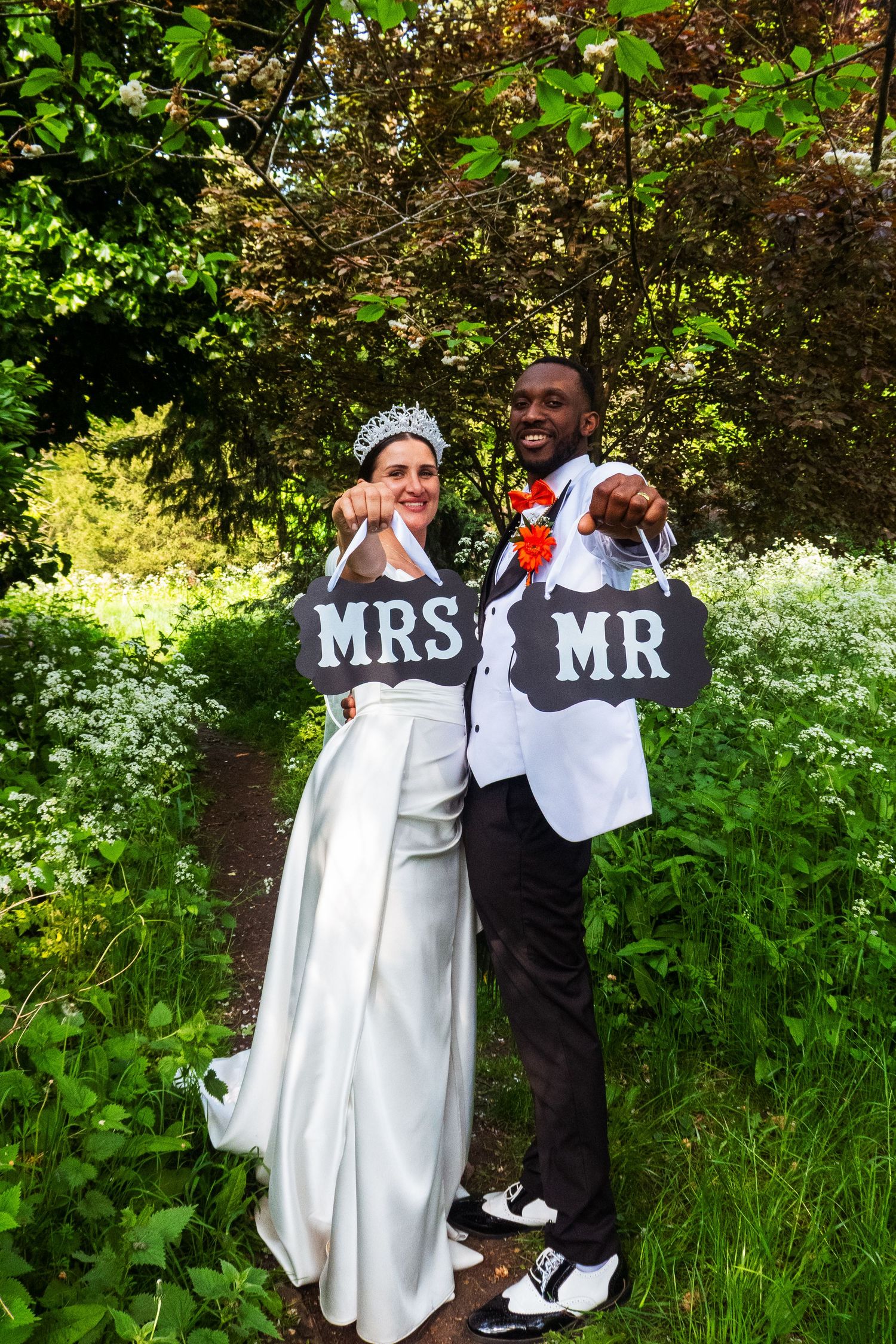 A bride and groom hold 'Mr' and 'Mrs' signs while posing in a lush garden setting.