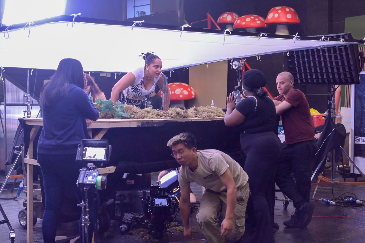 Film crew members work around table with red mushroom props visible in background.