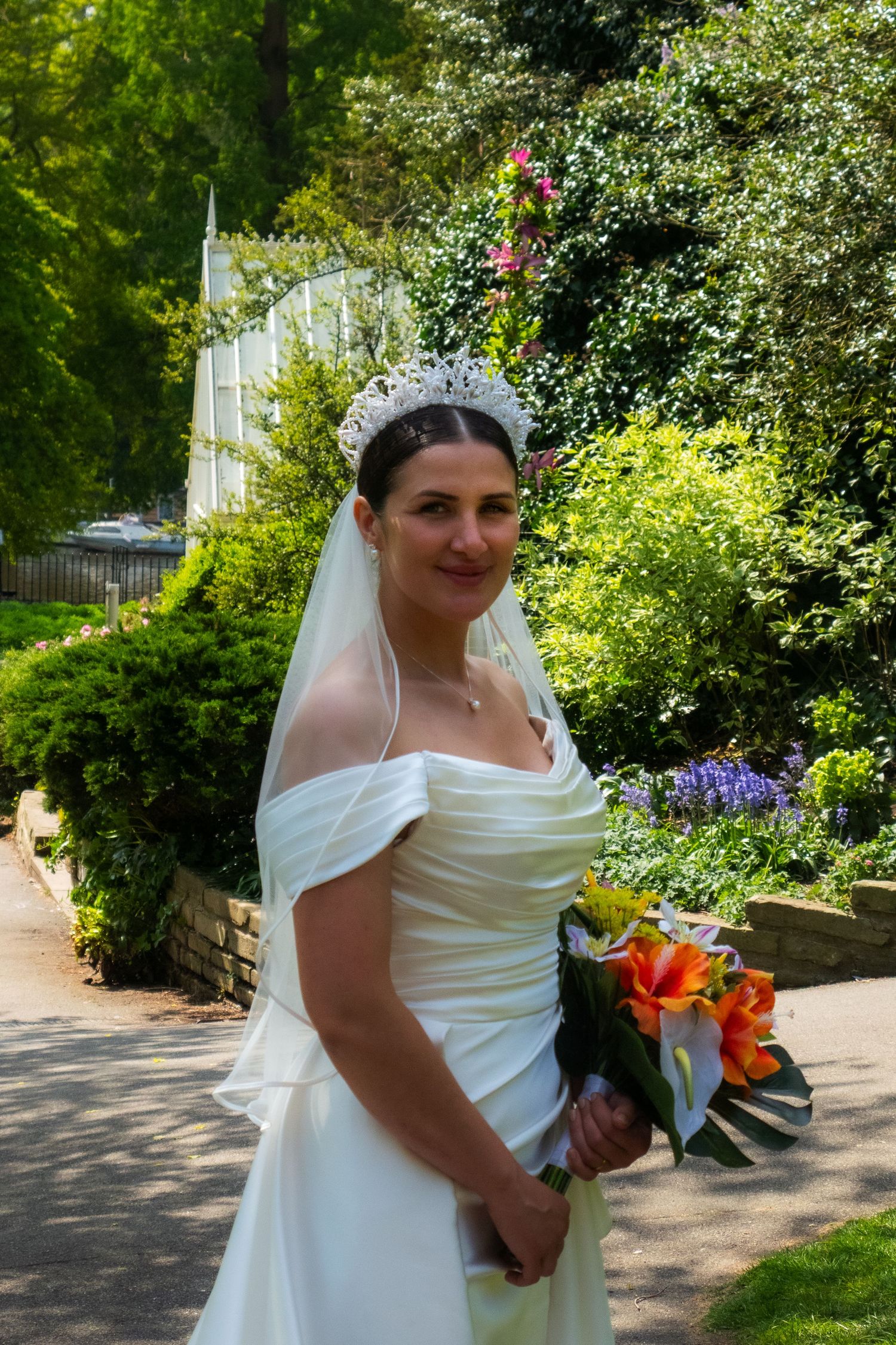A bride in an elegant off-shoulder white gown poses in a lush garden setting with colorful flowers.