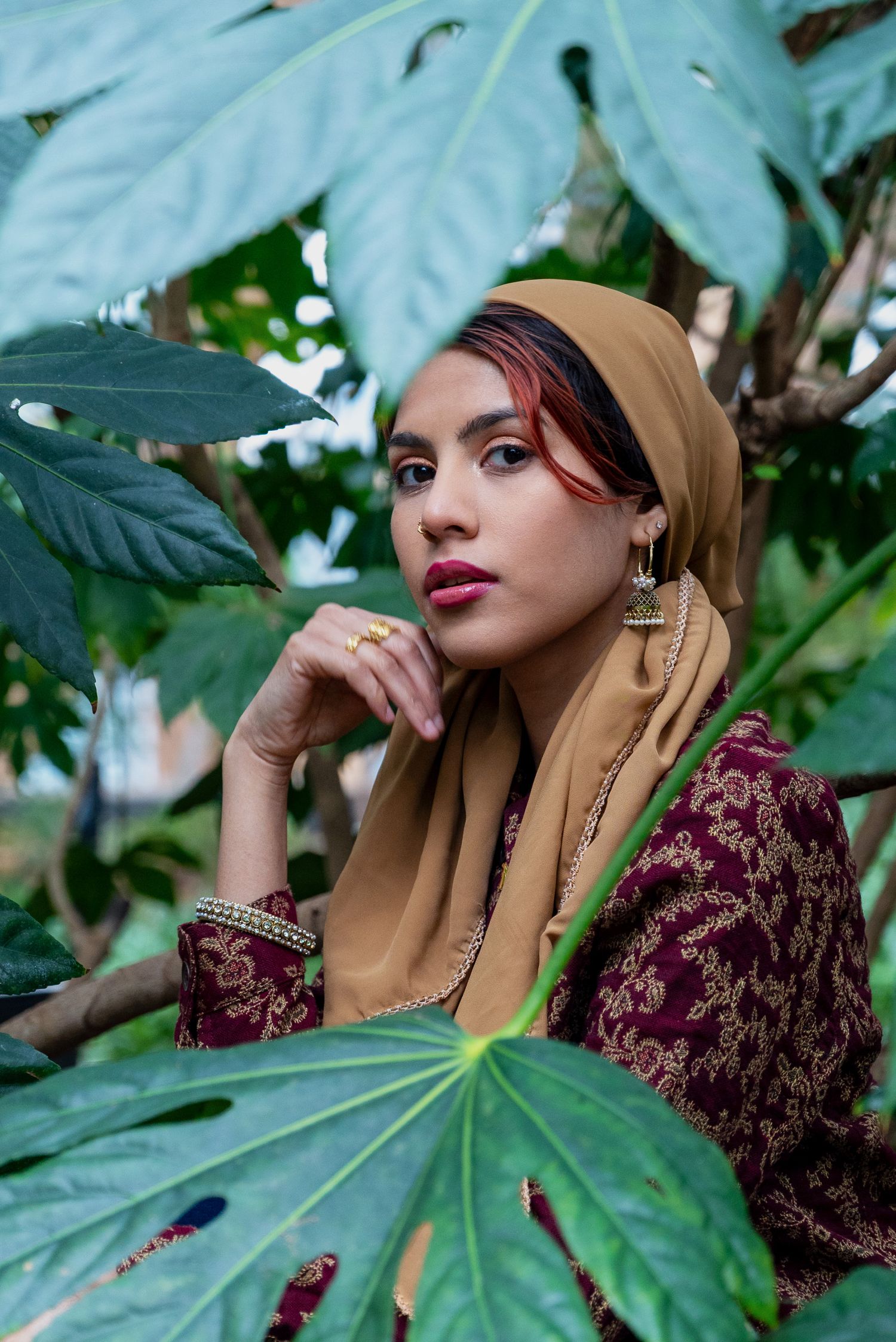 A woman in a tan hijab and burgundy embroidered dress poses thoughtfully among large tropical leaves outdoors.