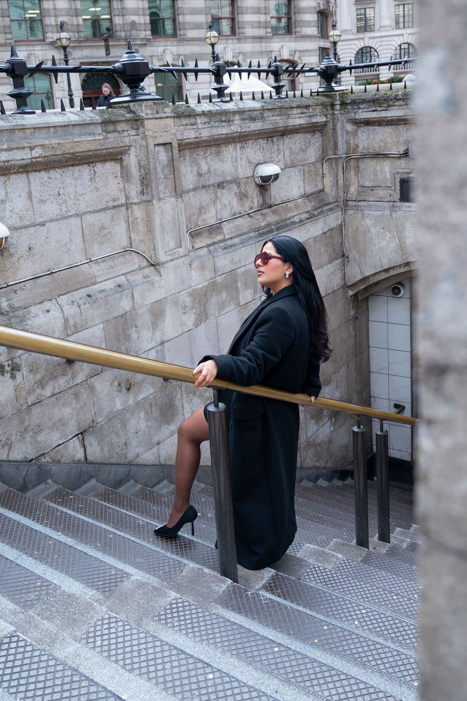 Person in black coat performs ballet stretch exercises on outdoor stone steps with metal handrail.