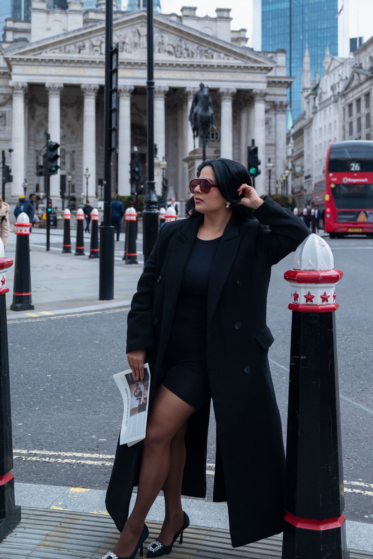 Fashionable woman in long black coat poses on London street with Royal Exchange and red double-decker bus in background.