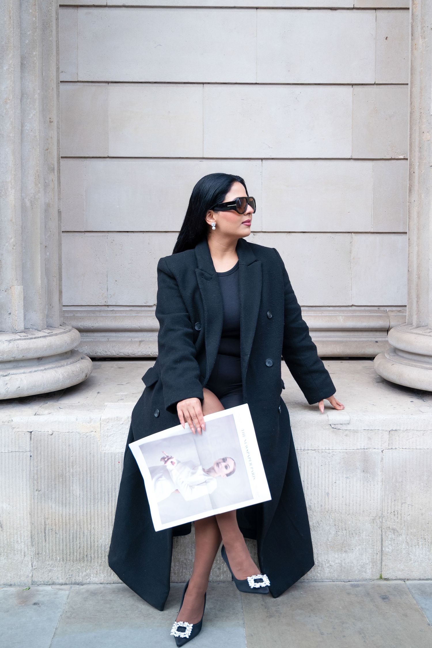 A person in a black blazer sits against a stone wall holding papers while posing in four different positions.