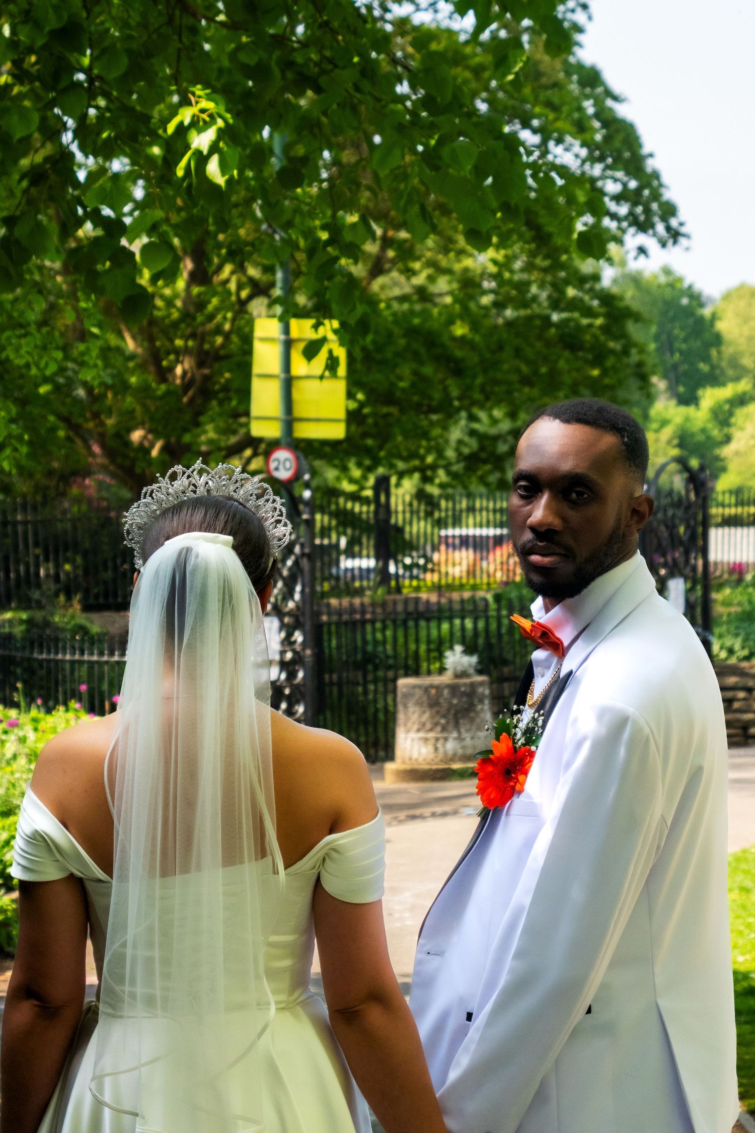 A wedding couple stands in a garden setting with lush greenery and iron fencing in the background.