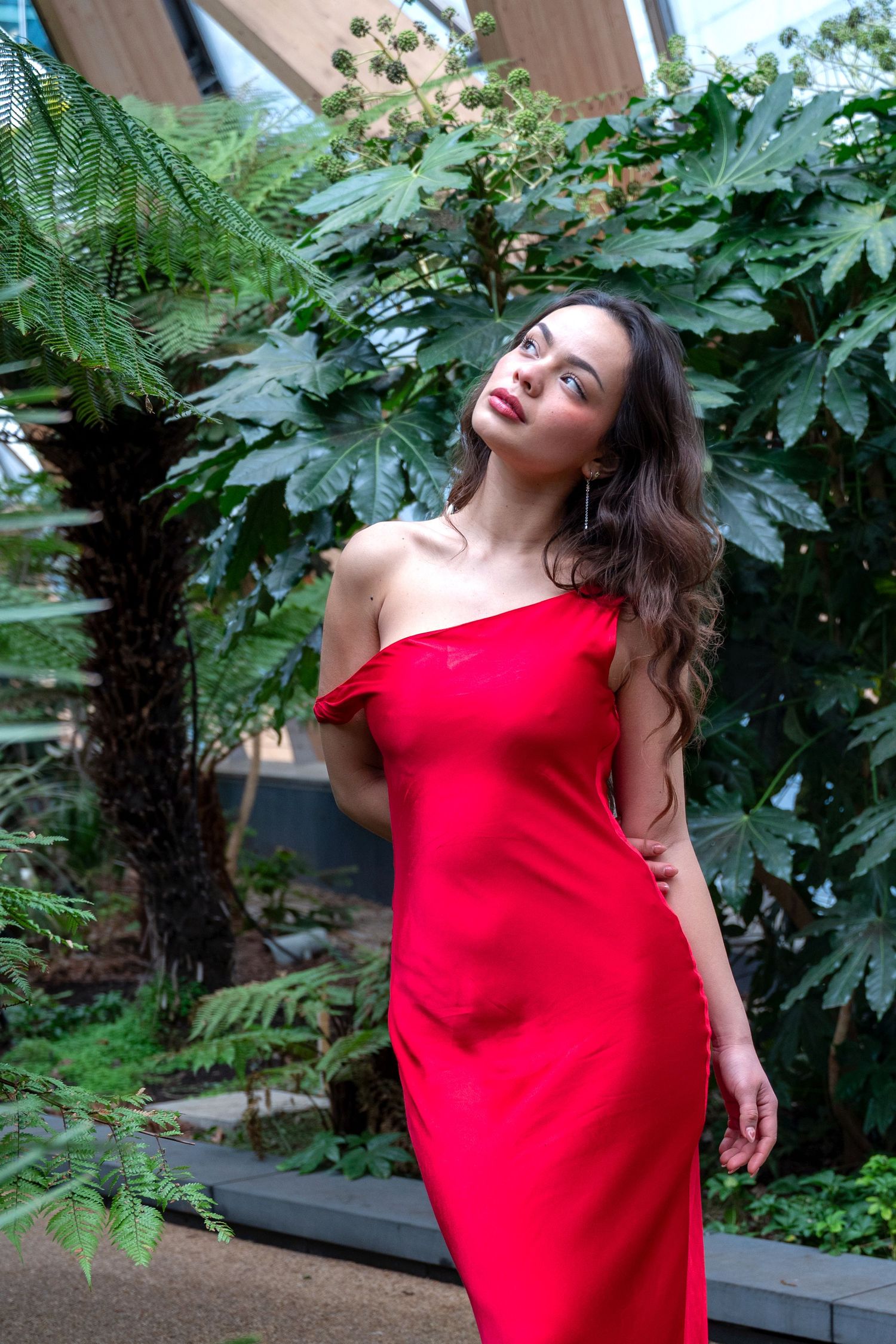 Woman in elegant red one-shoulder dress posing among lush tropical greenery and ferns in a botanical garden.