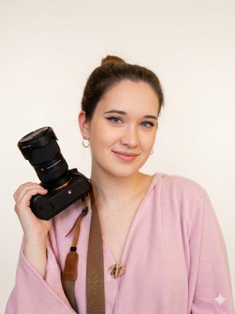 Young woman in pink cardigan holding a professional DSLR camera, smiling confidently against a white background.