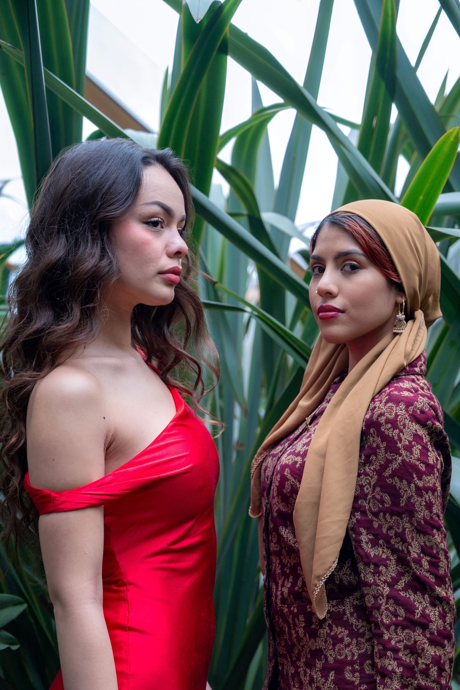Two women pose before tropical plants, one in a red off-shoulder dress, the other in a floral outfit with a tan hijab.