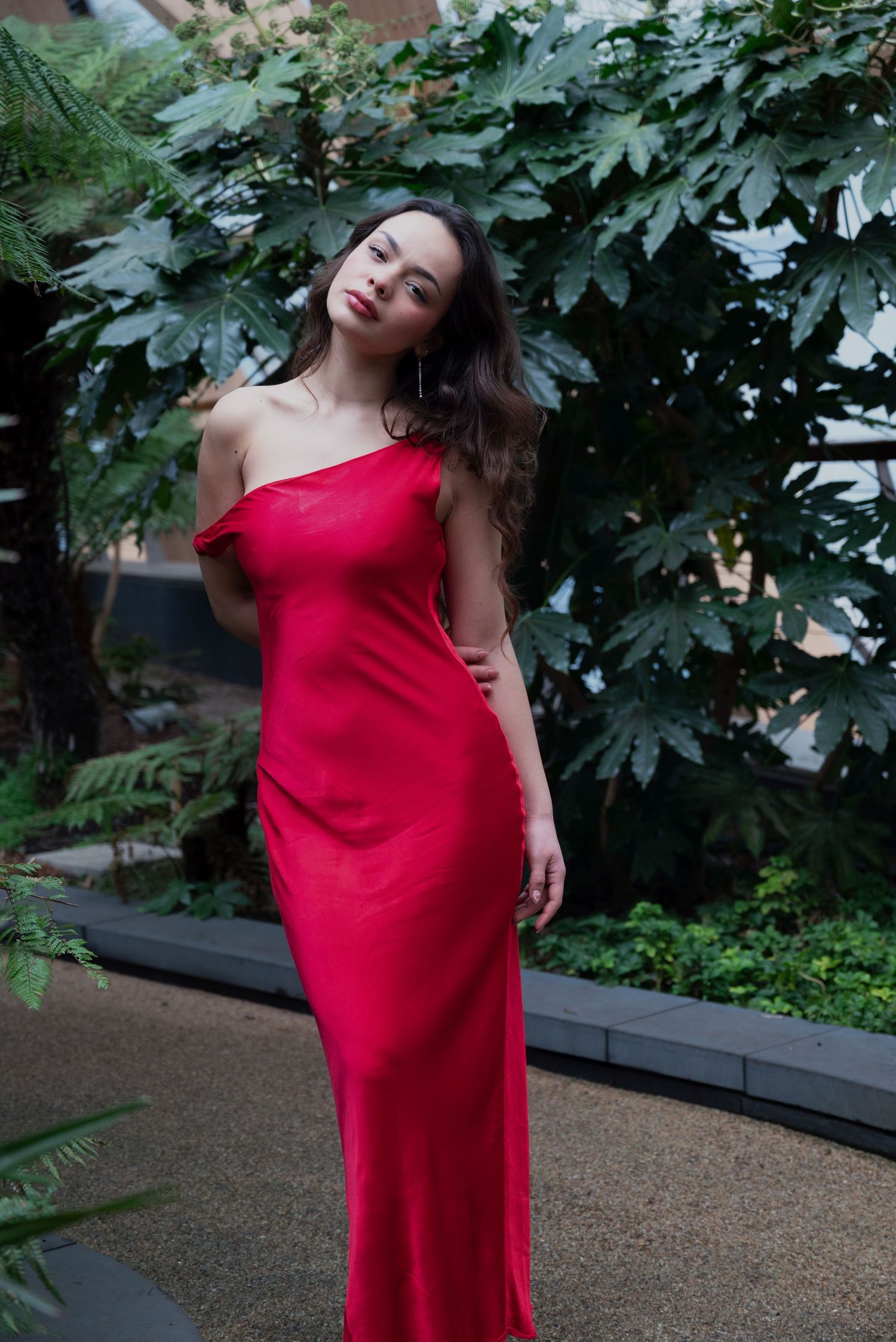 Woman in elegant red off-shoulder satin gown posing gracefully among lush tropical foliage.